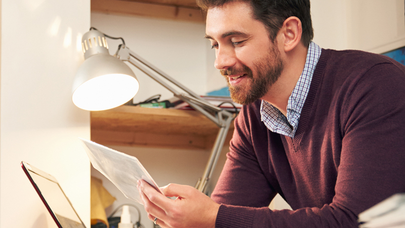 Man reviewing a document under a desk lamp in front of a laptop.